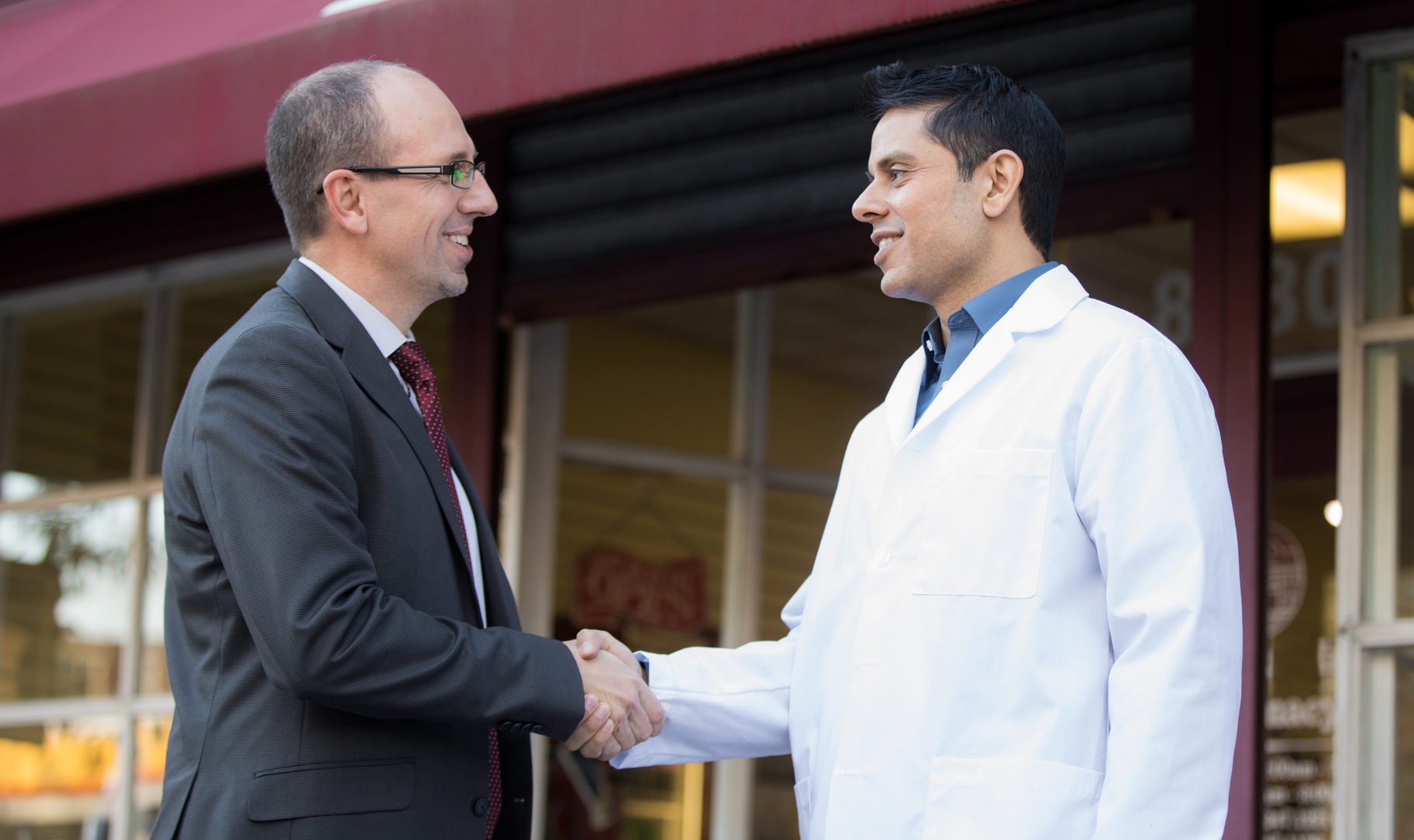 A young pharmacist shaking hands with an acquisition advisor in front of a pharmacy